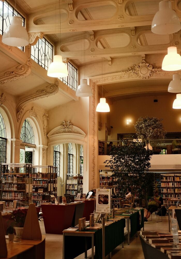 Interior grandioso de biblioteca com tetos altos, arquitetura ornamentada, grandes janelas, estantes de livros e &aacute;reas de leitura aconchegantes.
