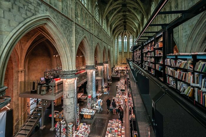 Grandiose interior de biblioteca com tetos abobadados, arcos de pedra e fileiras de estantes cheias de livros e visitantes navegando.