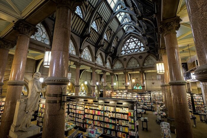 Interno grandioso di biblioteca con colonne imponenti, soffitti a volta e scaffali pieni di libri in un'ambientazione magica.
