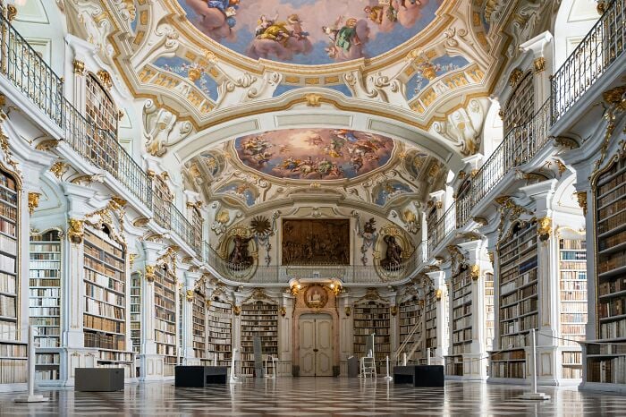 Interior grandioso de biblioteca com tetos ornamentados e estantes altas repletas de livros em uma atmosfera m&aacute;gica.