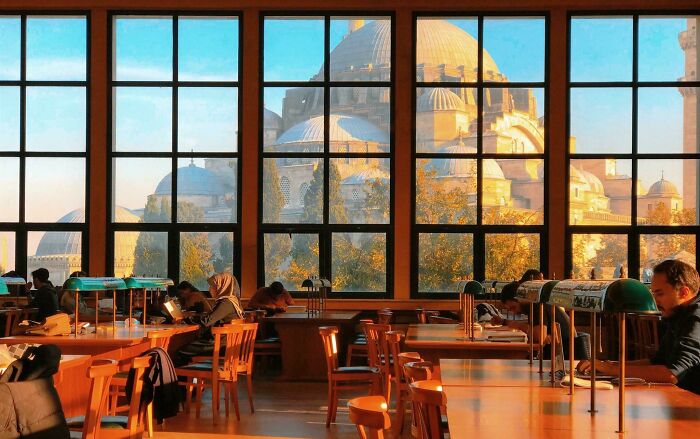 Cozy library interior with large windows showing grandiose architecture, and people reading and studying at wooden tables.